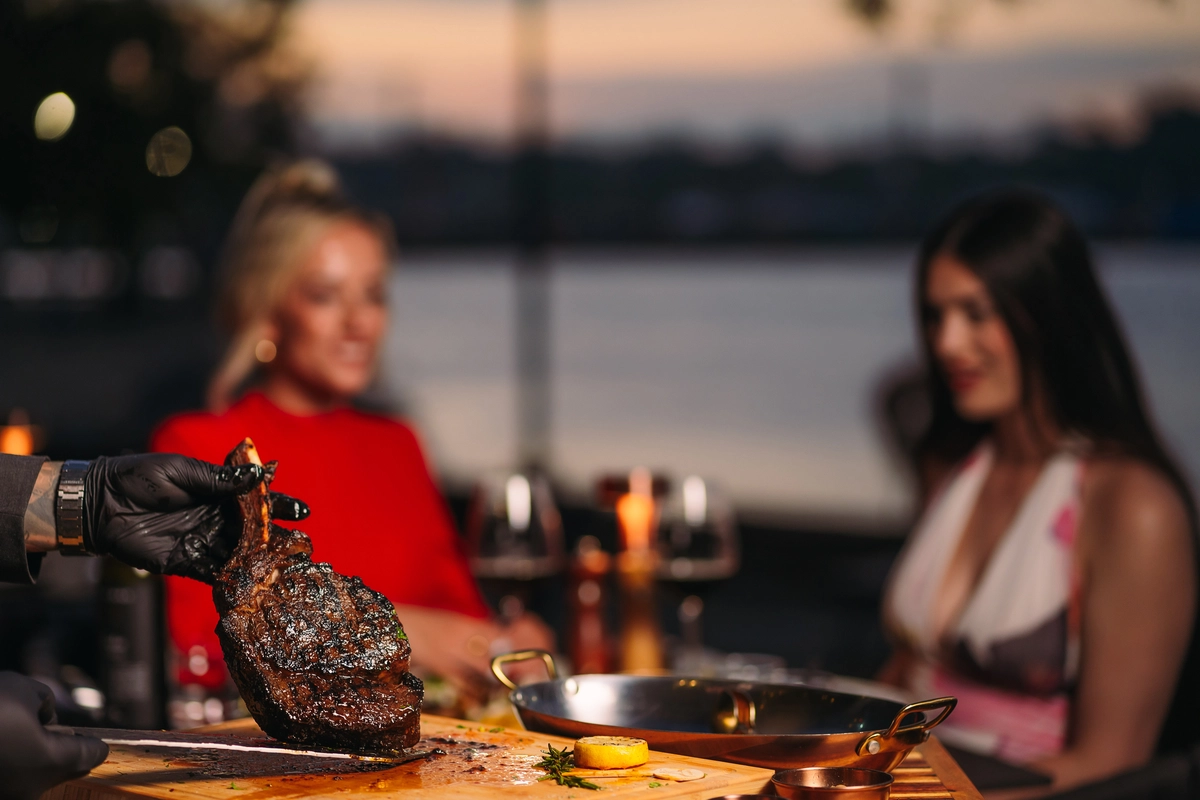A tomahawk carved at the table as two guests dine by the harbour at dusk